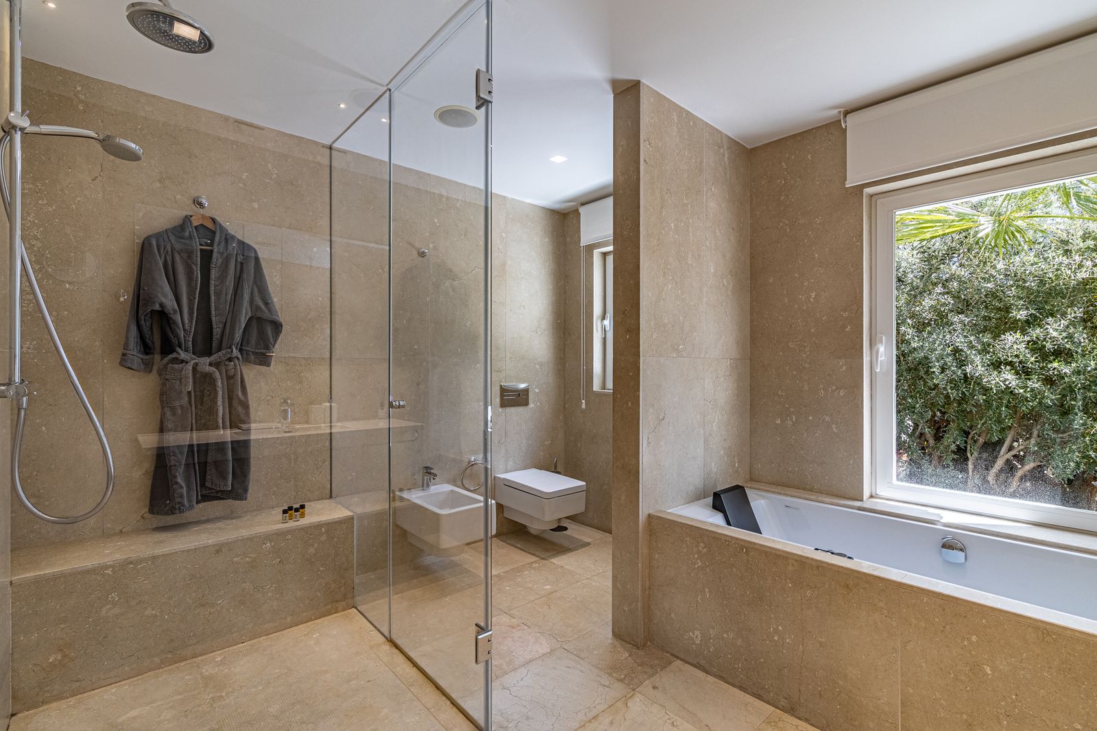 Beige stone bathroom with a glass-enclosed shower and a window overlooking greenery