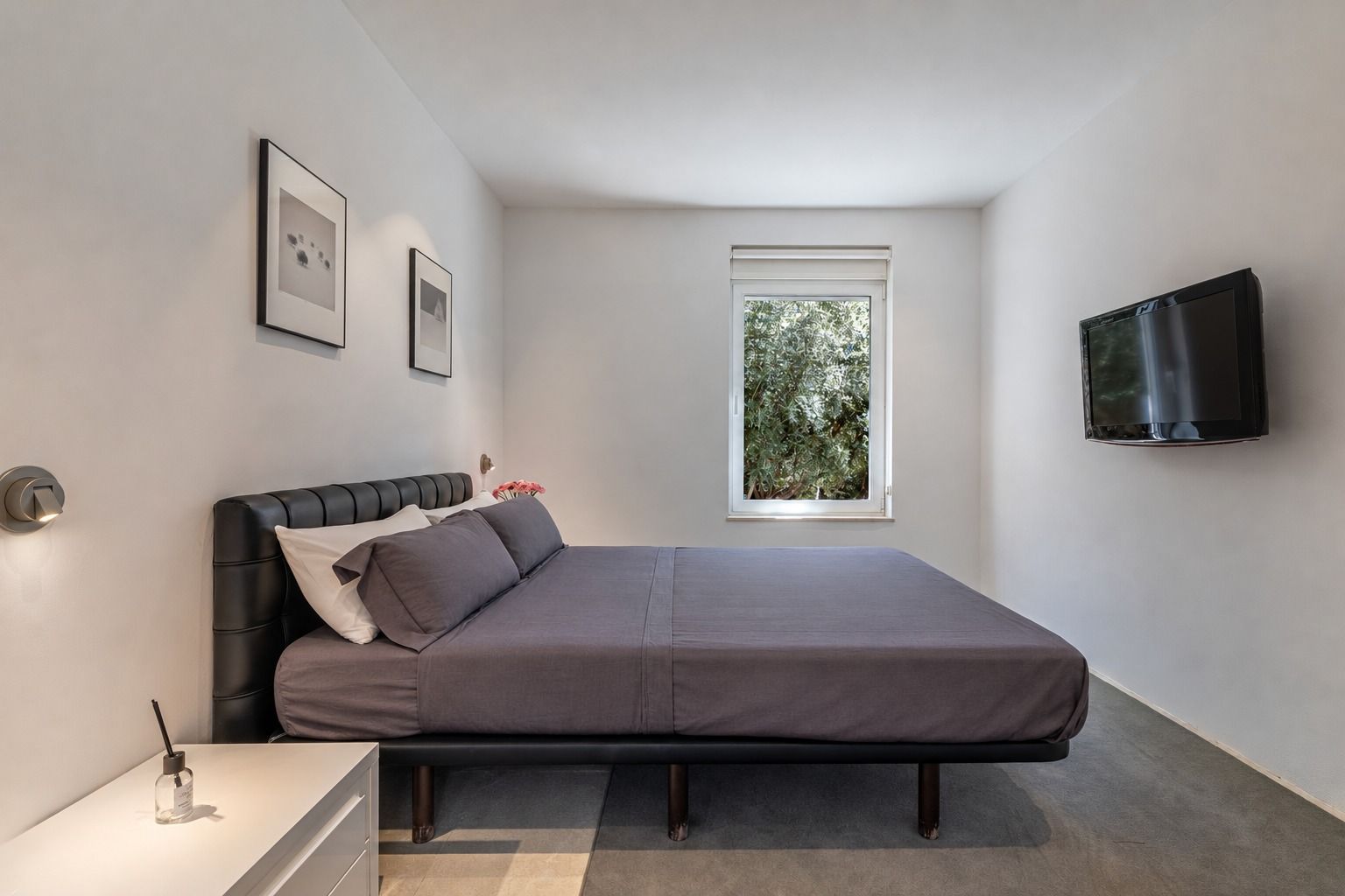 Bedroom with a double bed with a dark padded headboard, a wall TV and a window onto greenery
