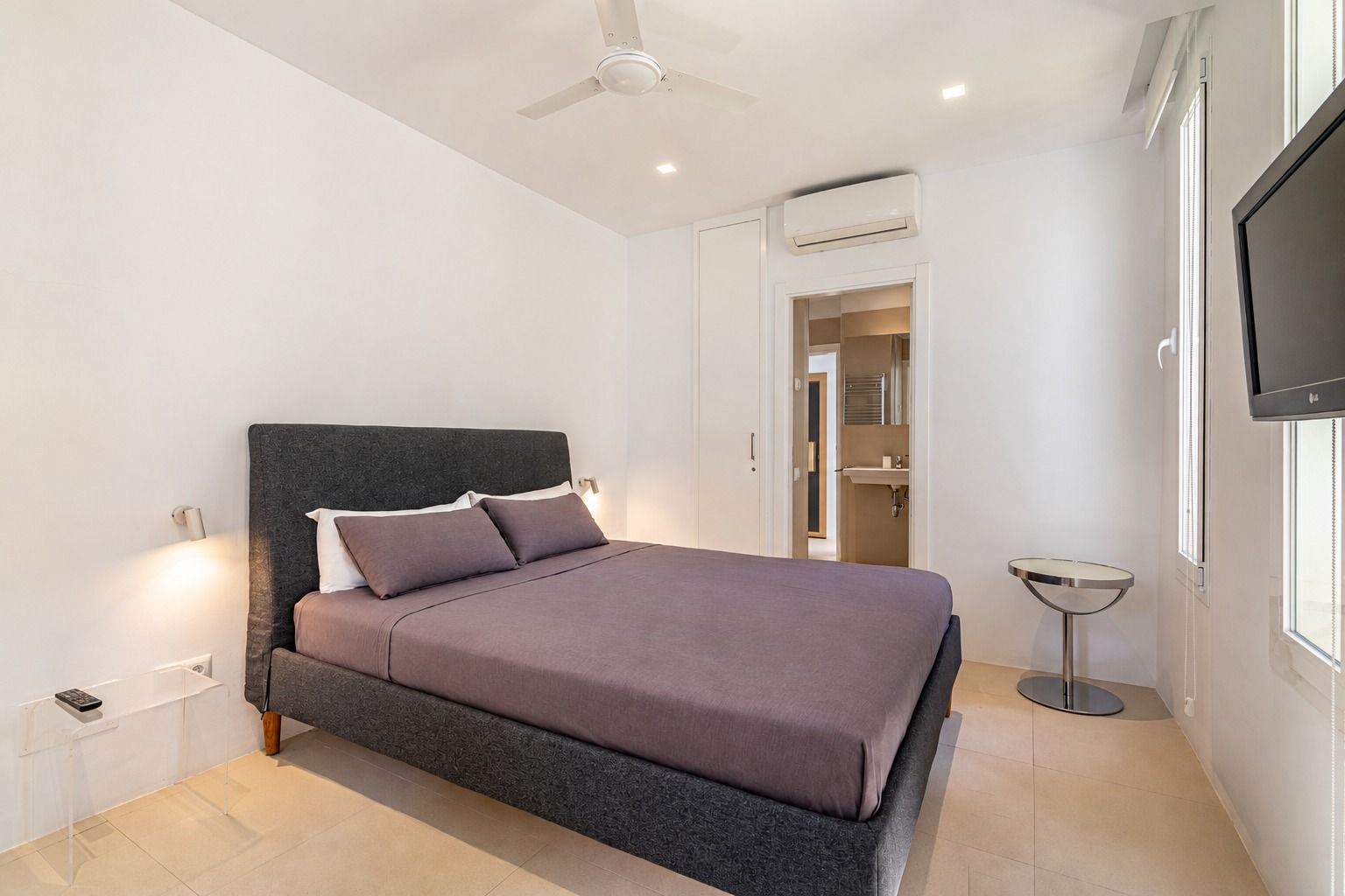 Compact bedroom with a dark grey fabric double bed, a ceiling fan and the en-suite bathroom door