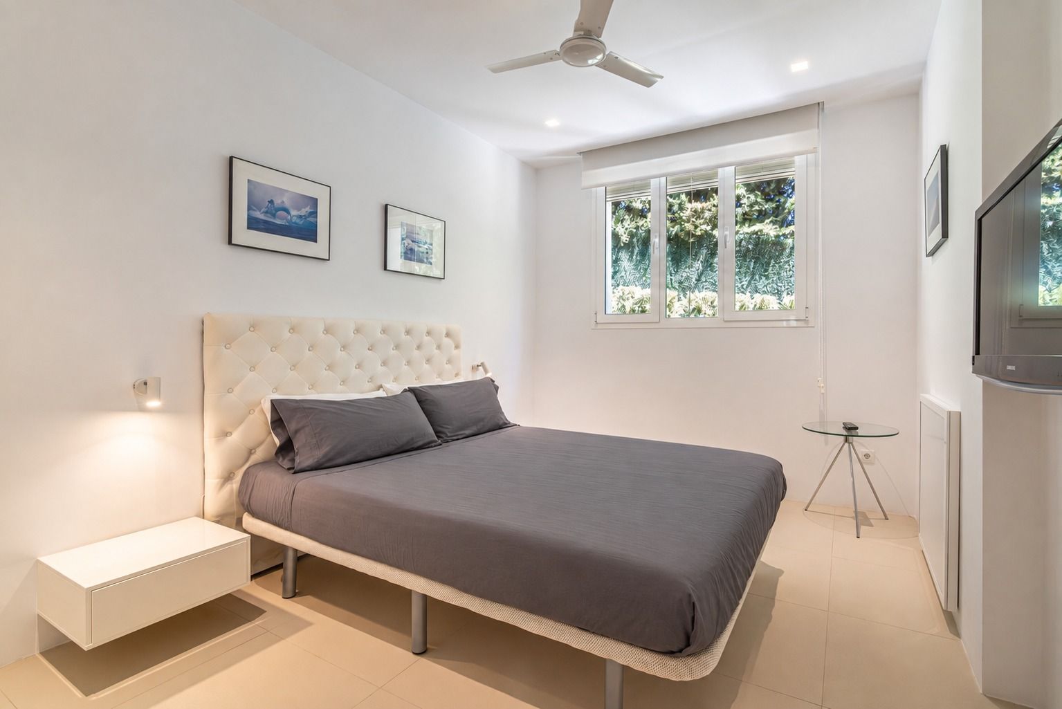 Bedroom with a white tufted headboard, a ceiling fan and a window onto the garden