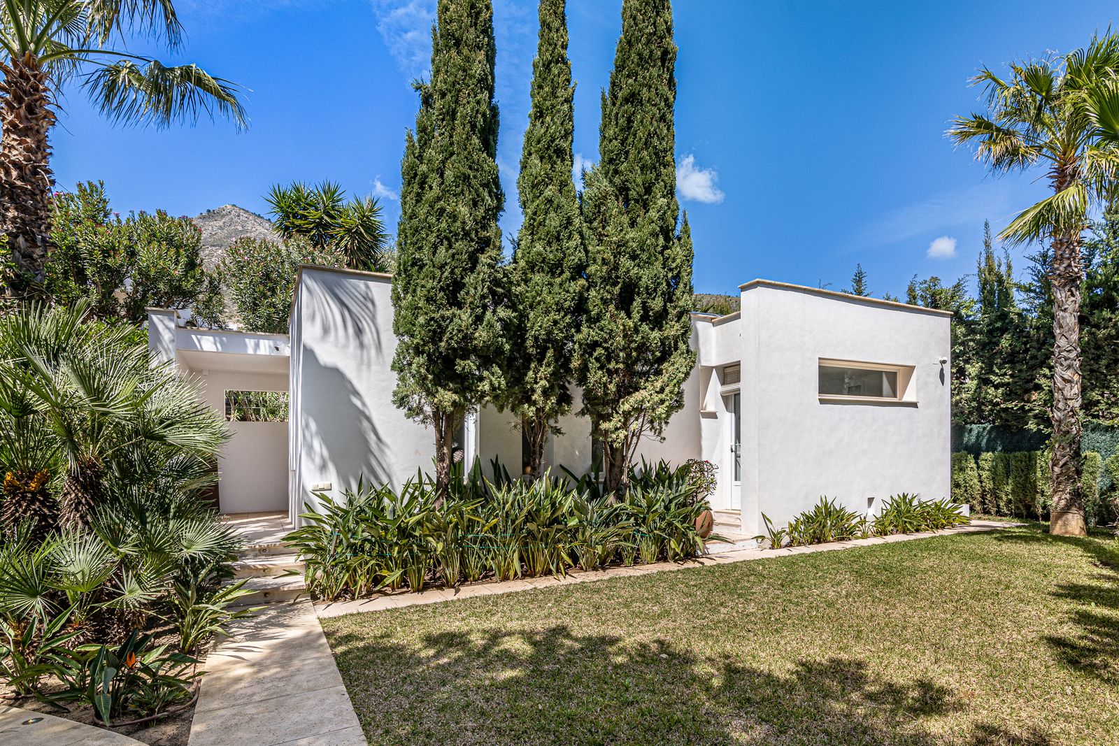 White guest house framed by cypress trees, with a lawn and mountains beyond