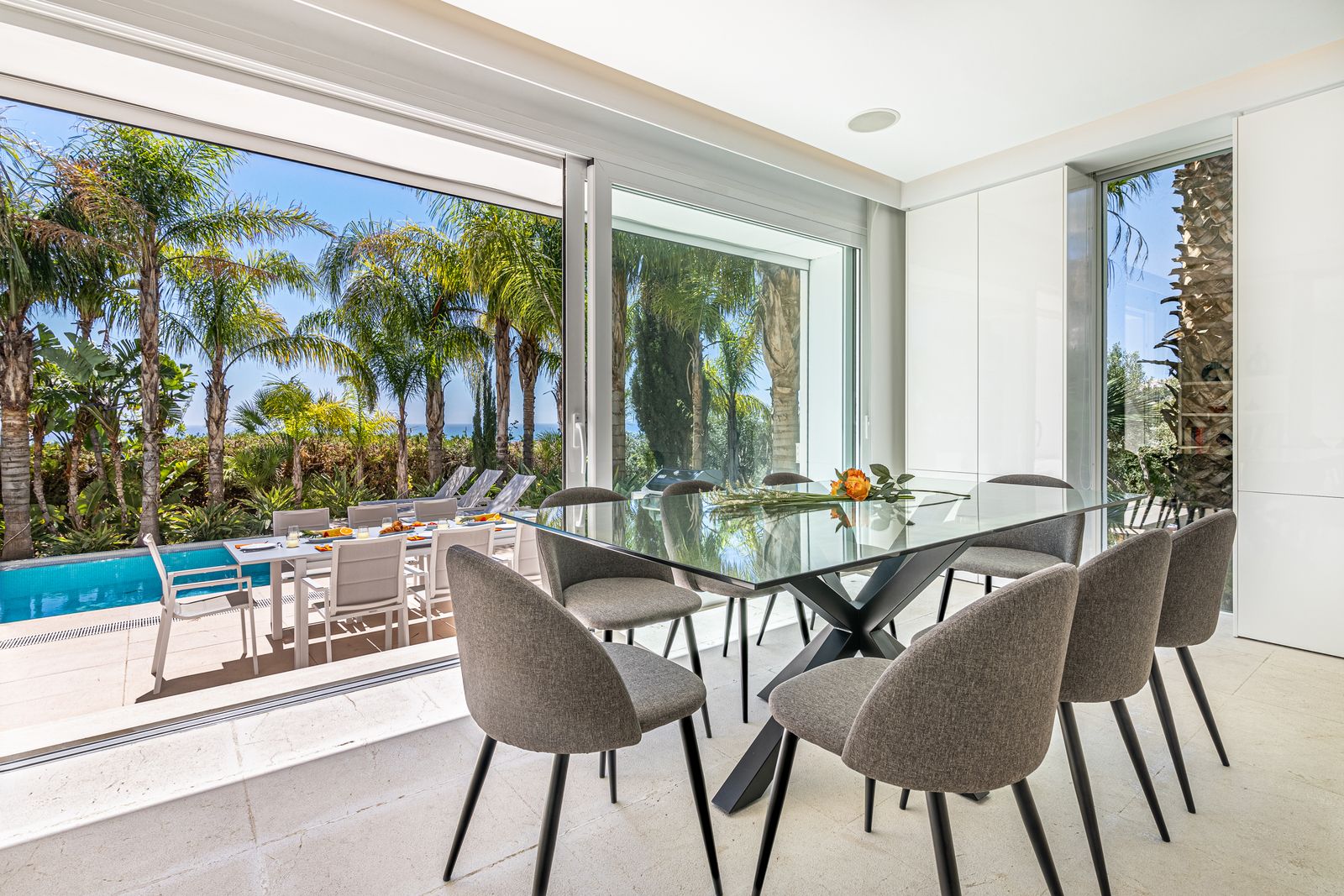Glass dining table and grey chairs overlooking the palm garden and pool