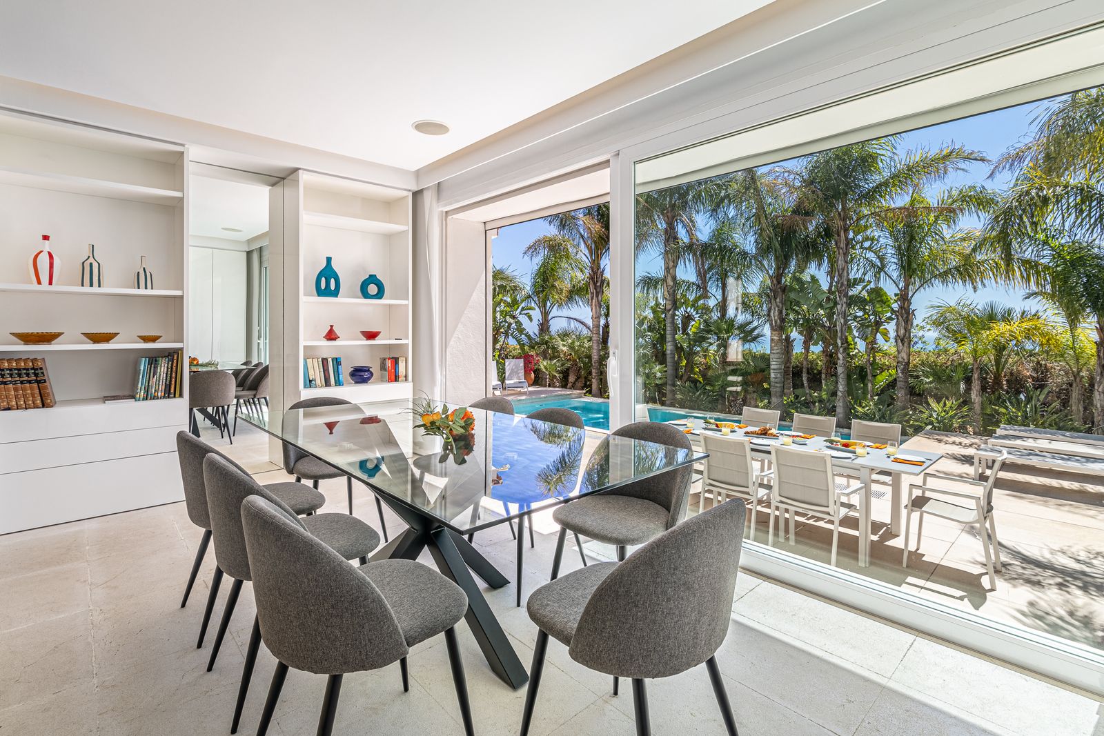 Glass dining table and chairs beside large windows overlooking the pool and palm trees